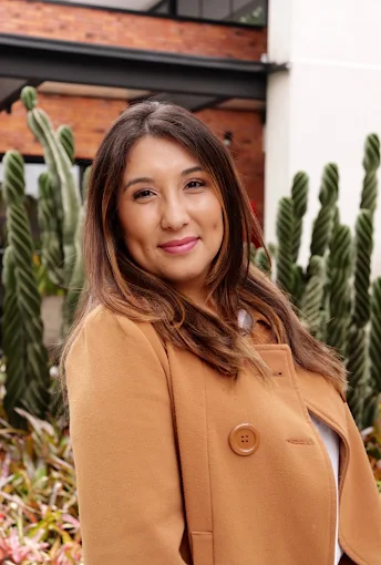 Retrato da psicóloga Flor Gonzalez sorrindo, de casaco caramelo, em frente a cactos
